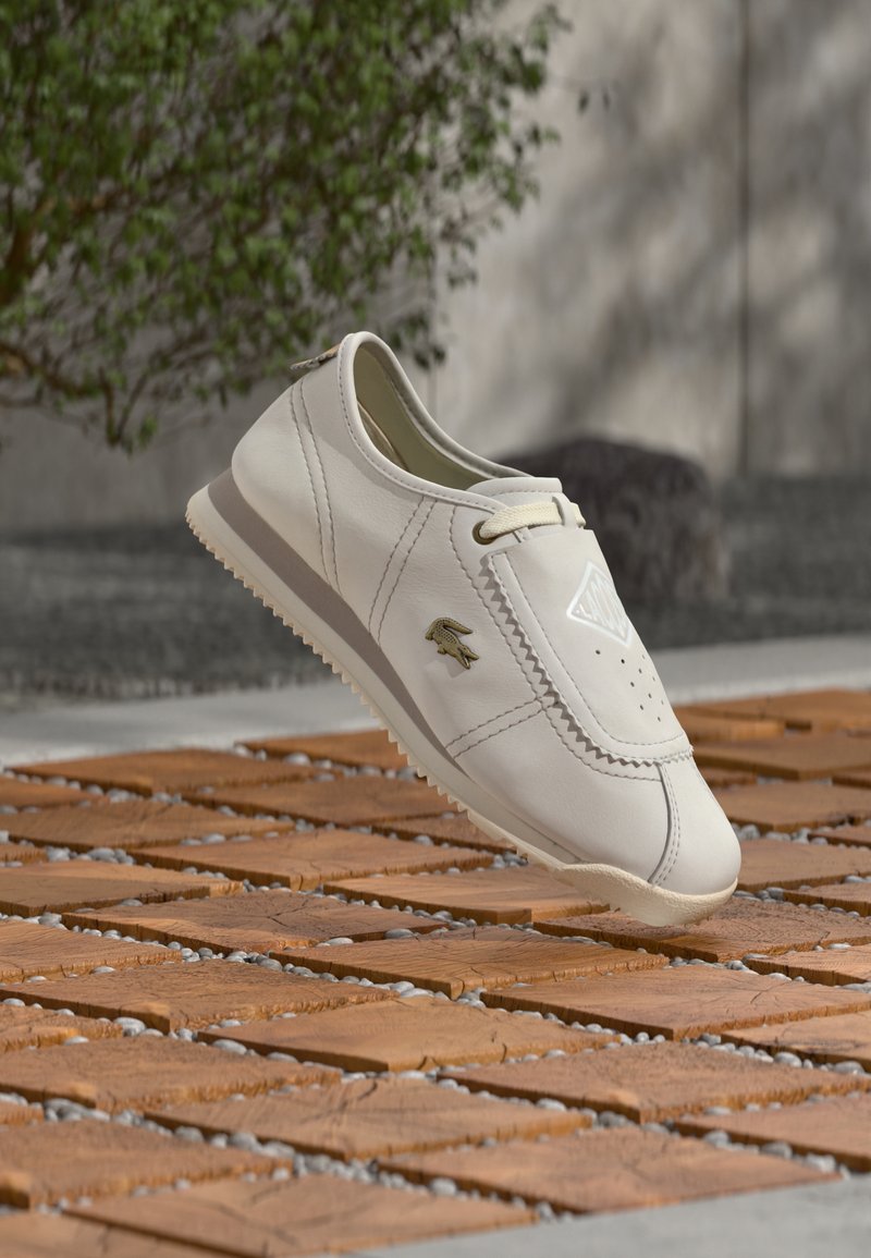 Baskets blanches légères en cuir avec détails perforés, accents beige et semelle en caoutchouc texturée, présentées au-dessus d'une surface en bois.