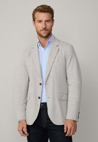 Ljusgrå blazer i mjukt tyg, med två knappar, två framfickor, bärs över en ljusblå skjorta med klassisk krage och denimjeans.
