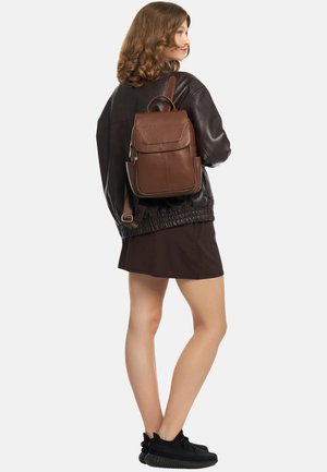 Bruine lederen rugzak met een gestructureerde afwerking, een voorflap, een ritsvak en verstelbare riemen, gedragen over een zwart bomberjack.