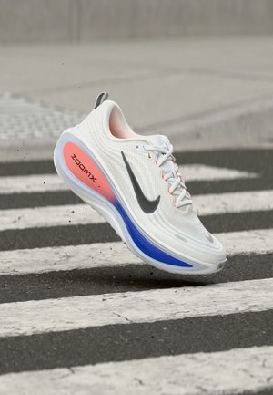 Chaussure de course Nike ZoomX blanche et grise avec semelle bleu et rose survolant un passage piéton sur l’asphalte.