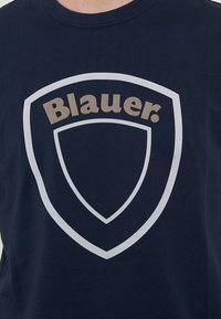 Mørkeblå T-shirt med et stort skjoldlogo med "Blauer." i brun og en hvid kant. Glat bomuldsstruktur, crew neck design.