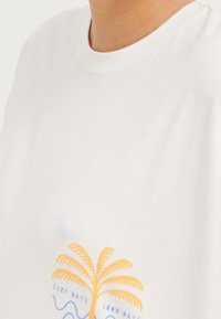 Camiseta blanca con un pequeño gráfico de una palmera naranja y texto azul que dice "Días de Surf" y "Buenos Días" cerca del área del pecho.