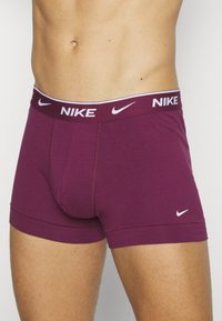 Burgundi színű Nike bokszerruha fehér logóval, puha anyagból, rugalmas derékpánttal és testhezálló fazonnal. Zárt sliccel és varrás nélküli lábnyílásokkal rendelkezik.