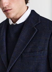 Blazer navy strutturato con colletto a punta e taschino sul petto, indossato sopra un maglione scuro, caratterizzato da sottili accenti marroni e un colletto a coste.