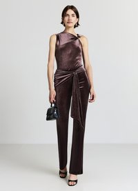 Tuta di velluto bordeaux con scollo asimmetrico, dettaglio annodato sulla spalla, parte frontale drappeggiata con fiocco, indossata con borsa nera e tacchi.