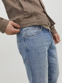Personne portant un jean bleu et une veste marron, ajustant le bas de la veste près de la taille avec une main.
