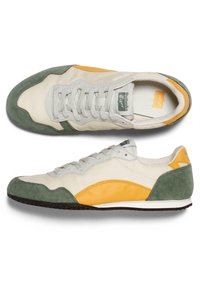 Vita och gröna mockasneakers med gula accenter. Har en texturerad tåhuvud, snören och en kontrasterande svart gummisul.