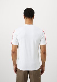 Λευκό T-shirt κοντό μανίκι με κόκκινες λεπτομέρειες κατά μήκος των ραφών στους ώμους. Φτιαγμένο από μαλακό βαμβακερό υλικό με κανονική εφαρμογή και στρογγυλή λαιμόκοψη.