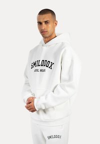 Sweatshirt à capuche blanche avec "SMILODOX" et "ATHL. WEAR" imprimés en noir. Comprend une poche avant et des poignets et ourlet côtelés.