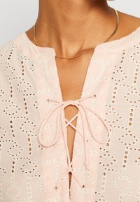 Gros plan sur une personne portant un chemisier en dentelle œillet rose pâle avec un nœud noué au niveau de l'encolure et un collier fin en chaîne dorée.