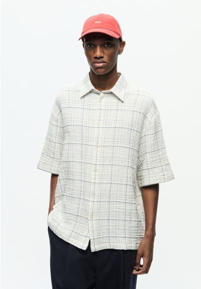 Chemise à manches courtes en tissu léger crème avec un motif à carreaux bleus, fermeture à boutons et col étalé. Casquette rouge avec logo brodé.