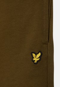 Olivegrüner Baumwollstoff mit glatter Textur, der ein gesticktes schwarzes und gelbes Adlerlogo auf der unteren Seite zeigt.