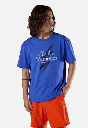 Młody mężczyzna z kręconymi włosami, ubrany w niebieską koszulkę "Trail Memories" i jaskrawo pomarańczowe spodenki, stoi z jedną ręką w kieszeni, patrząc w prawo.