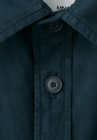 Gros plan sur le col d'une chemise bleu foncé boutonnée, avec des coutures visibles et un bouton noir fermé au centre.