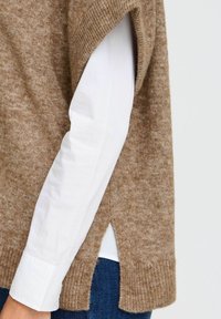 Brun strikket sweater med en blød tekstur, korte ærmer, sideslids og lagdelt med en hvid skjorte med knapper under.