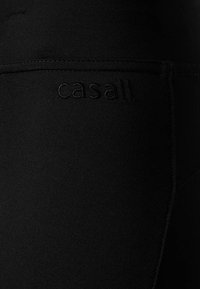 Czarne legginsy z elastycznego materiału mają matowe wykończenie, elastyczny pas i wytłoczone logo "casall." z boku.