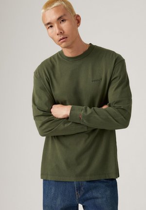 Olive green long-sleeve shirt in cotton with a crew neck, featuring a small black logo on the chest and ribbed cuffs. Worn with blue jeans.