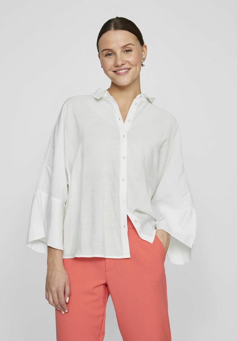 Camicia bianca oversized con bottoni sul davanti, colletto a punta e maniche ampie, abbinata a pantaloni rosa a sigaretta. Tessuto dalla texture liscia.
