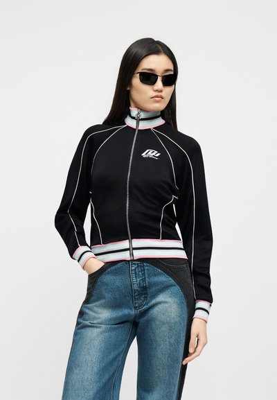 Γυναίκα με μακριά σκούρα μαλλιά που φοράει μαύρα γυαλιά ηλίου, μαύρο μπουφάν με φερμουάρ και παστέλ ρίγες, καθώς και τζιν δύο αποχρώσεων, στέκεται μπροστά σε λευκό φόντο.