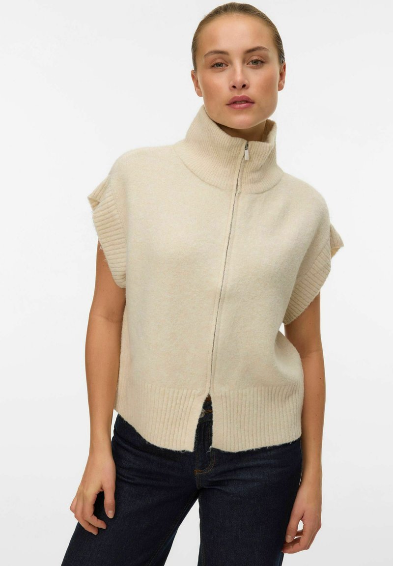 Maglione senza maniche beige lavorato a maglia con colletto alto e zip frontale, presenta dettagli a coste all'orlo e al colletto, texture morbida.