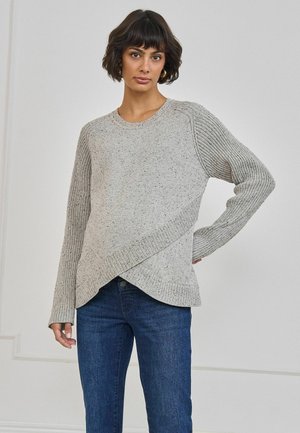 Femme aux cheveux courts et foncés portant un pull en maille gris clair avec un motif côtelé diagonal et un jean bleu, debout contre un mur uni.