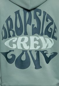 Vêtement à capuche avec un texte audacieux et incurvé "Dropside Crew Gouve" en bleu foncé et bleu clair sur un tissu vert pâle.