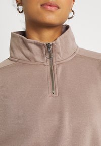 Jasnobrązowa bluza z suwakiem przy szyi i wysokim kołnierzem, wyposażona w srebrny zamek błyskawiczny z pierścieniowym uchwytem. Miękka w dotyku i o casualowym designie.