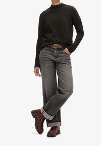 Pull en maille brun foncé, jean large gris et bottes de randonnée brunes. Le pull a un col montant ; le jean est retroussé à l'ourlet.