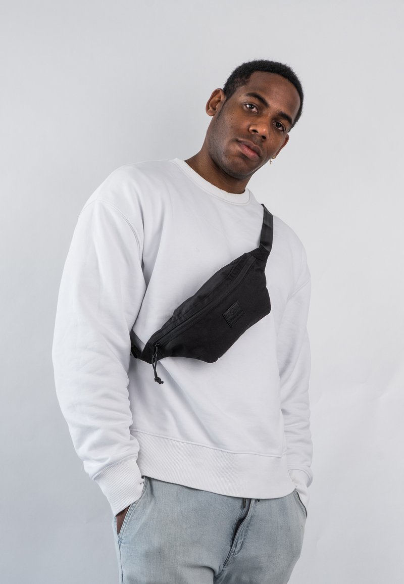 Banane en tissu noir portée en bandoulière ; elle dispose d'une fermeture éclair, d'une bandoulière réglable et d'un discret logo. Sweatshirt gris clair et jeans.