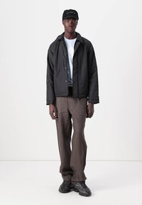Black insulated jacket, light blue t-shirt, brown wide-leg pants, and black sneakers; model wears a black cap with logo.