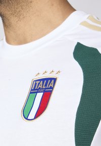 Hvid sportstrøje med et broderet italiensk flaglogo, gyldne accenter og grønne teksturerede skulderpaneler.