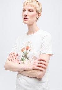 Άτομο με κοντά ξανθά μαλλιά που φοράει λευκό μπλουζάκι με πορτοκαλί τυπωμένα τριαντάφυλλα, σταυρωμένα χέρια και ασημένια σκουλαρίκια κρίκοι σε λευκό φόντο.