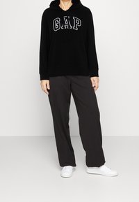 Sudadera negra con capucha con la palabra "GAP" impresa en blanco, combinada con pantalones grises holgados y zapatillas blancas sobre un fondo sencillo.