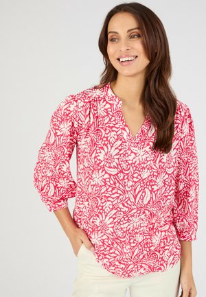 Mujer sonriendo, lleva una blusa de patrón floral roja y blanca con mangas 3/4, combinada con pantalones de color claro, mano en el bolsillo.