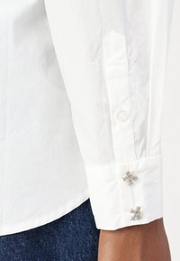 Poignet de chemise blanche à manches longues avec deux boutons de manchette argentés en forme de nœud, porté avec un jean denim bleu foncé.