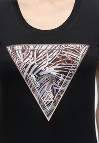 Μαύρο πουκάμισο με μεγάλο τριγωνικό λογότυπο Guess με ρίγες ζέβρας, στρας και κείμενο: "GUESS USA WASHED JEANS."