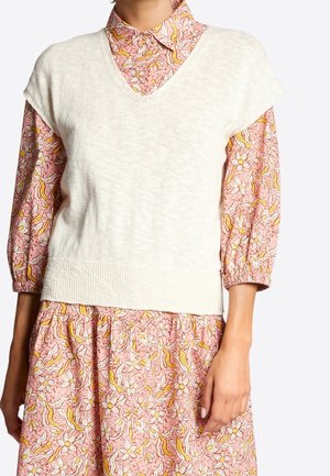 Chaleco de punto color crema con diseño de escote en V, usado sobre una blusa rosa con estampado que presenta detalles florales y mangas abullonadas.