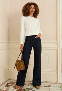 Blusa bianca a maniche lunghe con colletto in pizzo, abbinata a jeans a gamba larga blu scuro e una borsa con coulisse in suede marrone. Il modello indossa sandali marroni.