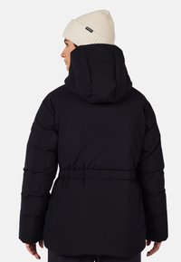 Person iført en sort hættetrøje vinterjakke og en off-white strikket hue, set fra ryggen mod en ensfarvet baggrund.