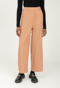 Kvinde iført beige vide bukser, sort langærmet top, sorte loafers med guldkæde-detalje, stående mod hvid baggrund.