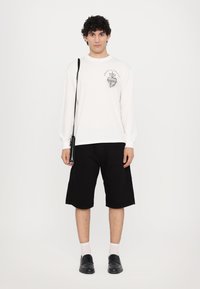 Cămașă albă cu mâneci lungi, cu un design grafic, pantaloni scurți negri, pantofi slip-on negri și o geantă mică pe umăr, tot neagră. Fundal simplu.