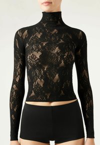 Haut en dentelle noire à manches longues avec un col haut, arborant des motifs floraux. Associé à un short noir ajusté, soulignant une silhouette élancée.