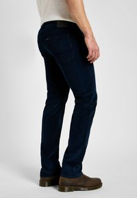 Jeans blu scuro in denim, con taglio dritto, cuciture sottili e tasche posteriori. Indossati con stivali marroni textured.