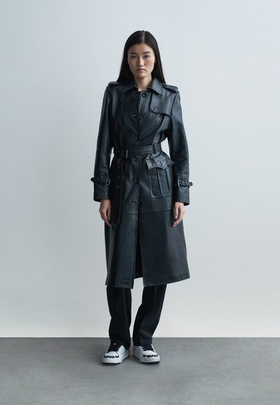 Trench in pelle nera con vita a cintura, grande tasca e chiusura con bottoni. Abbinato a pantaloni neri e sneakers bianche. Texture liscia.