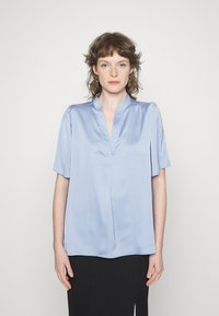 Blusa azul-claro de manga curta em tecido de satin suave, com uma pala frontal parcial e detalhes suaves com franzido nos ombros.
