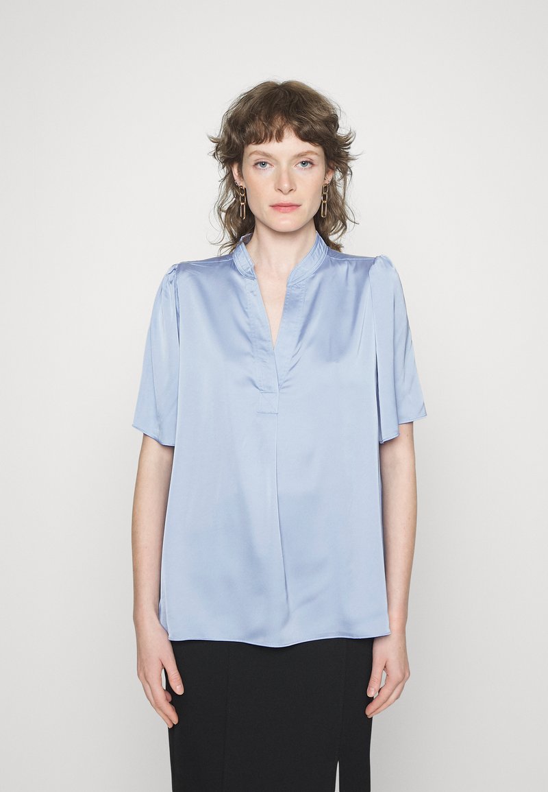 Blusa azul-claro de manga curta em tecido de satin suave, com uma pala frontal parcial e detalhes suaves com franzido nos ombros.