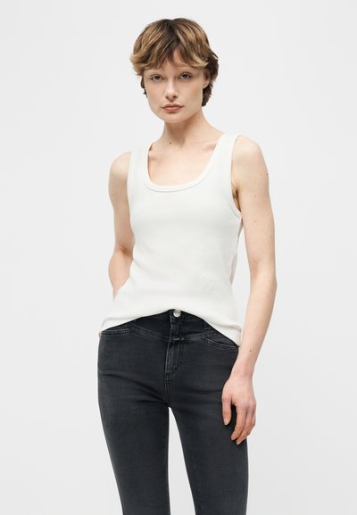 Νεαρό άτομο με κοντά μαλλιά, φορώντας λευκό αμάνικο τοπ και σκούρο γκρι τζιν, μπροστά σε απλό φόντο.