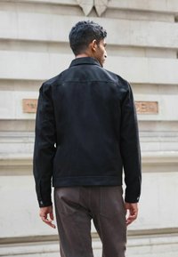 Schwarze Jeansjacke mit klassischem Kragen, schlichter Gestaltung und Knopfmanschetten, kombiniert mit gedeckten braunen Hosen. Rückansicht gezeigt.