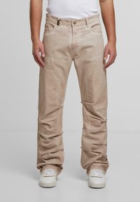 Mies, który ma na sobie jasnobeżowe luźne spodnie z bocznymi i przednimi kieszeniami, w połączeniu z białymi trampkami i białą koszulką bez rękawów.