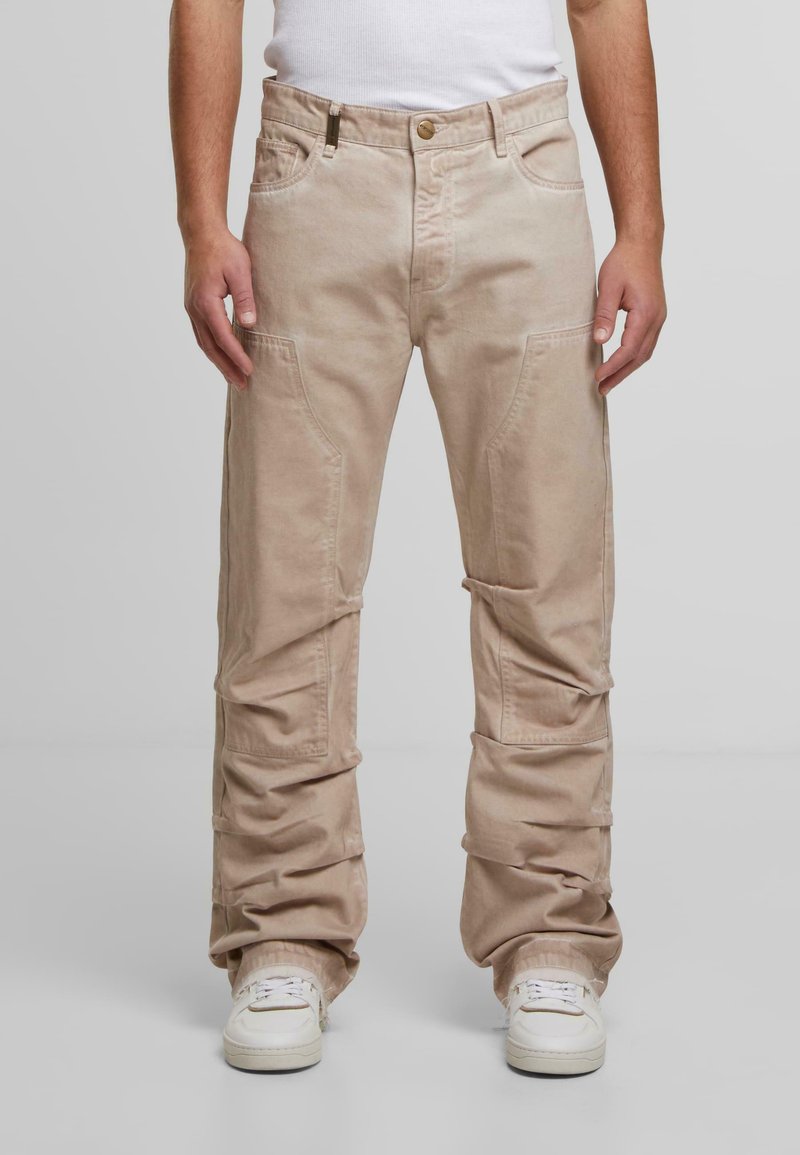 Mies, który ma na sobie jasnobeżowe luźne spodnie z bocznymi i przednimi kieszeniami, w połączeniu z białymi trampkami i białą koszulką bez rękawów.